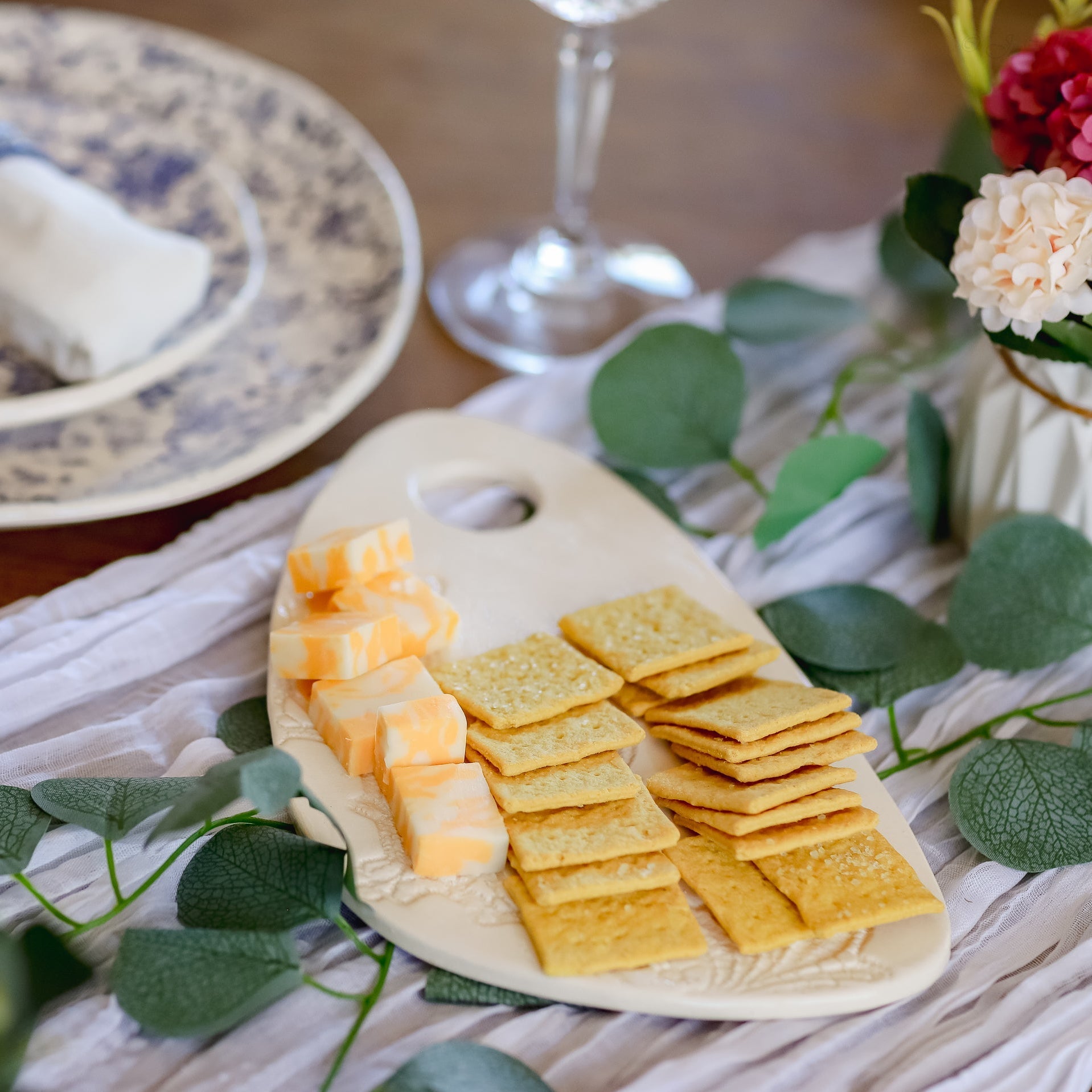 Platter of cheese and crackers on a table with a glass and flowers in the background