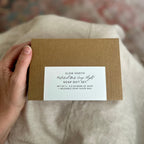 Hand holding a brown cardboard soap gift set box with a label on a soft fabric background