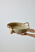 Handwoven basket with decorative handles and tassels held by a person on a light gray background