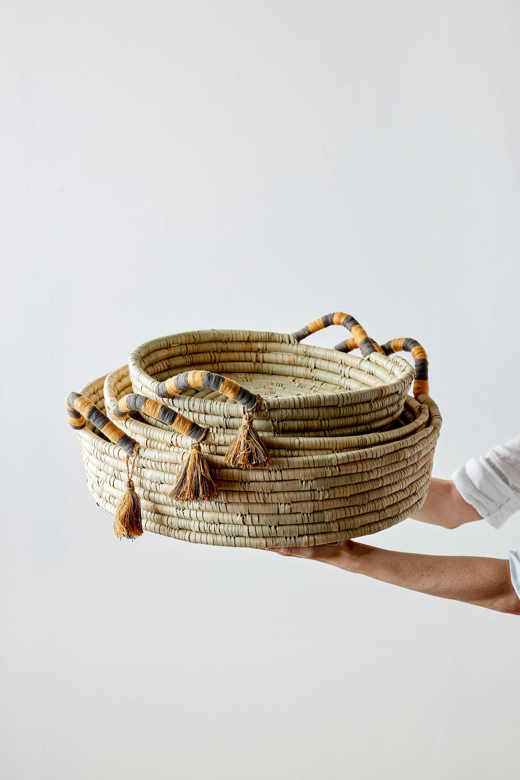 Person holding a stack of woven baskets with decorative tassels on a light background
