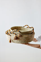 Person holding a stack of woven baskets with decorative tassels on a light background