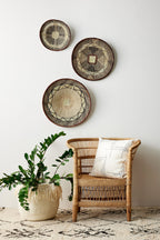 Wicker chair with a pillow next to a plant, with decorative woven plates on the wall.