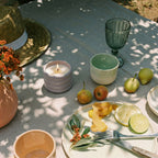 Outdoor setting with a table featuring a candle, fruit, and cutlery on a light surface.