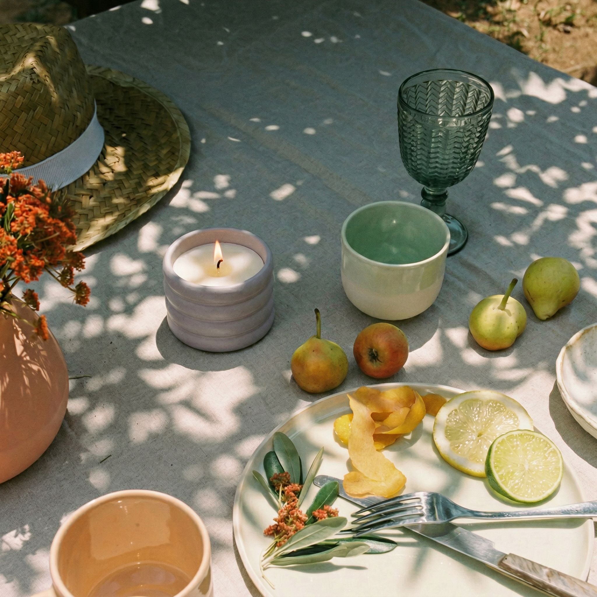Outdoor setting with a table featuring a candle, fruit, and cutlery on a light surface.