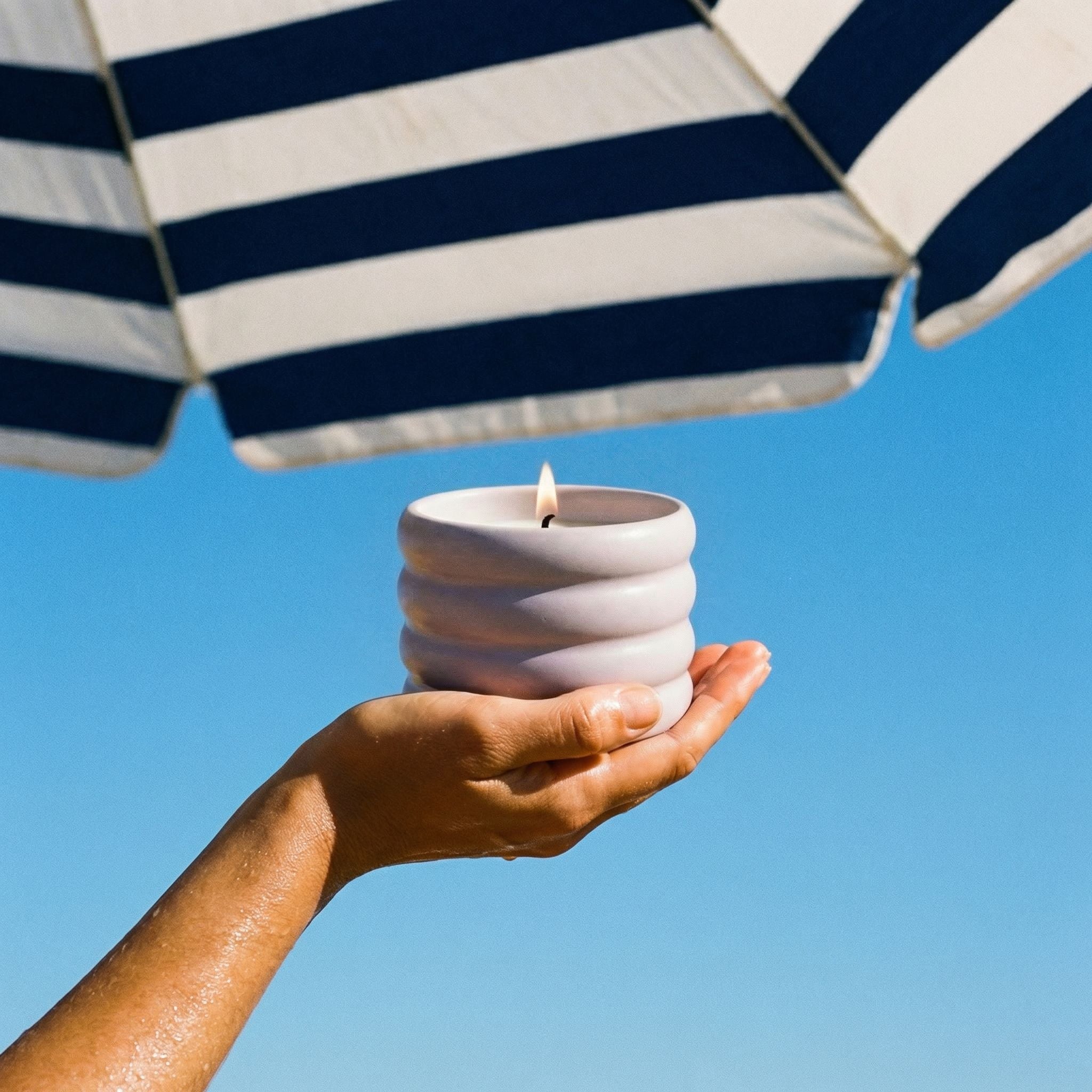Hand holding a white candle under a blue and white striped umbrella against a clear blue sky.