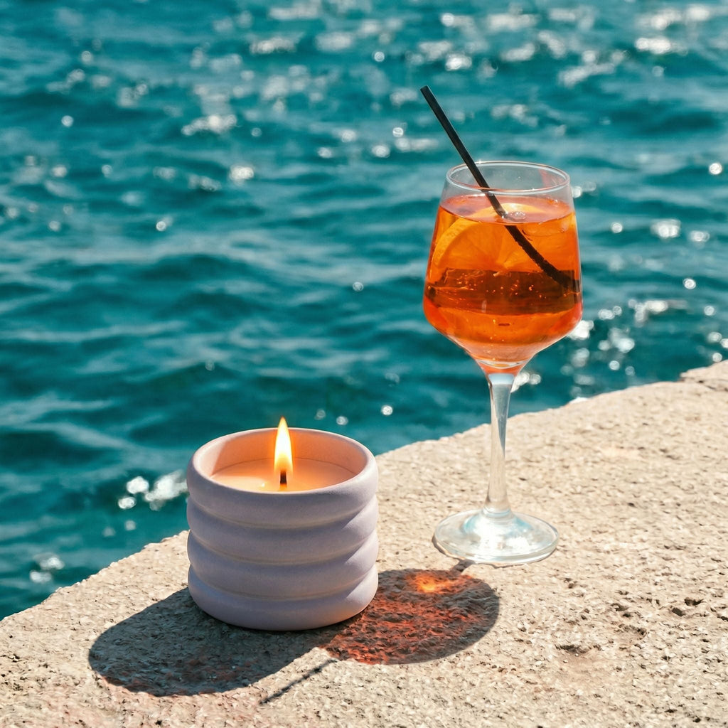 Glass of orange cocktail with a straw and lit candle on a ledge by the sea.