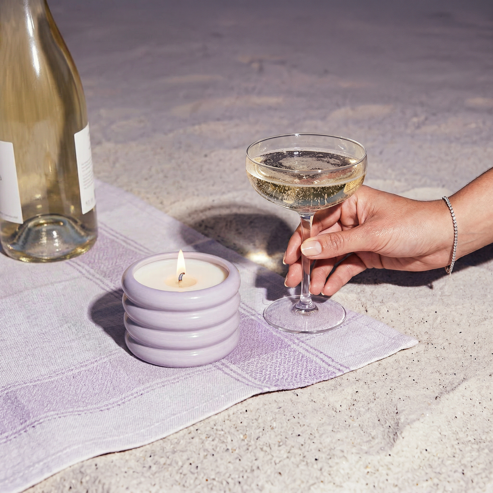 Hand holding a glass of champagne next to a lit candle and a wine bottle on a textured surface