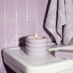 Candle in a decorative holder on a bathroom counter with purple tiles and a towel in the background.