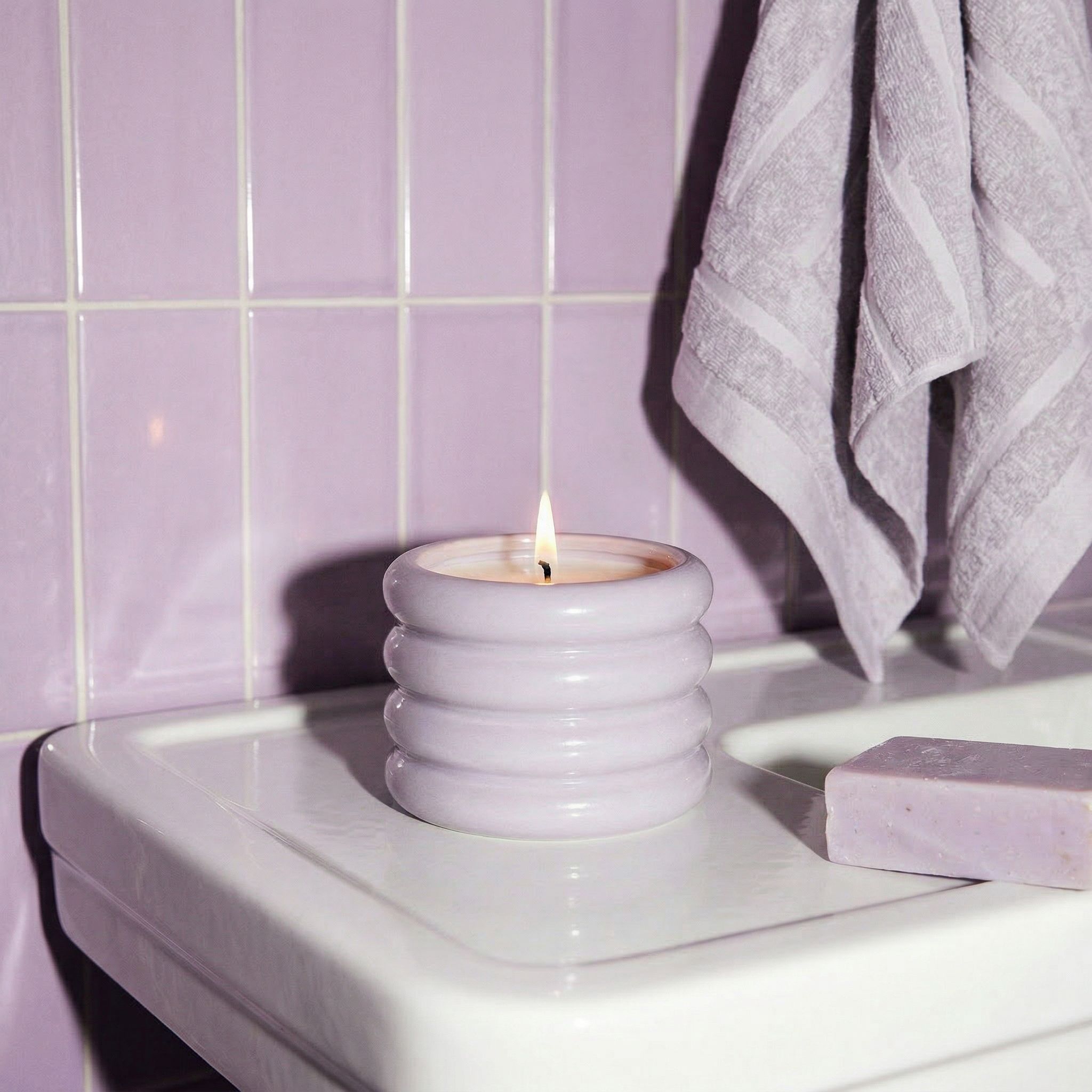 Candle in a decorative holder on a bathroom counter with purple tiles and a towel in the background.