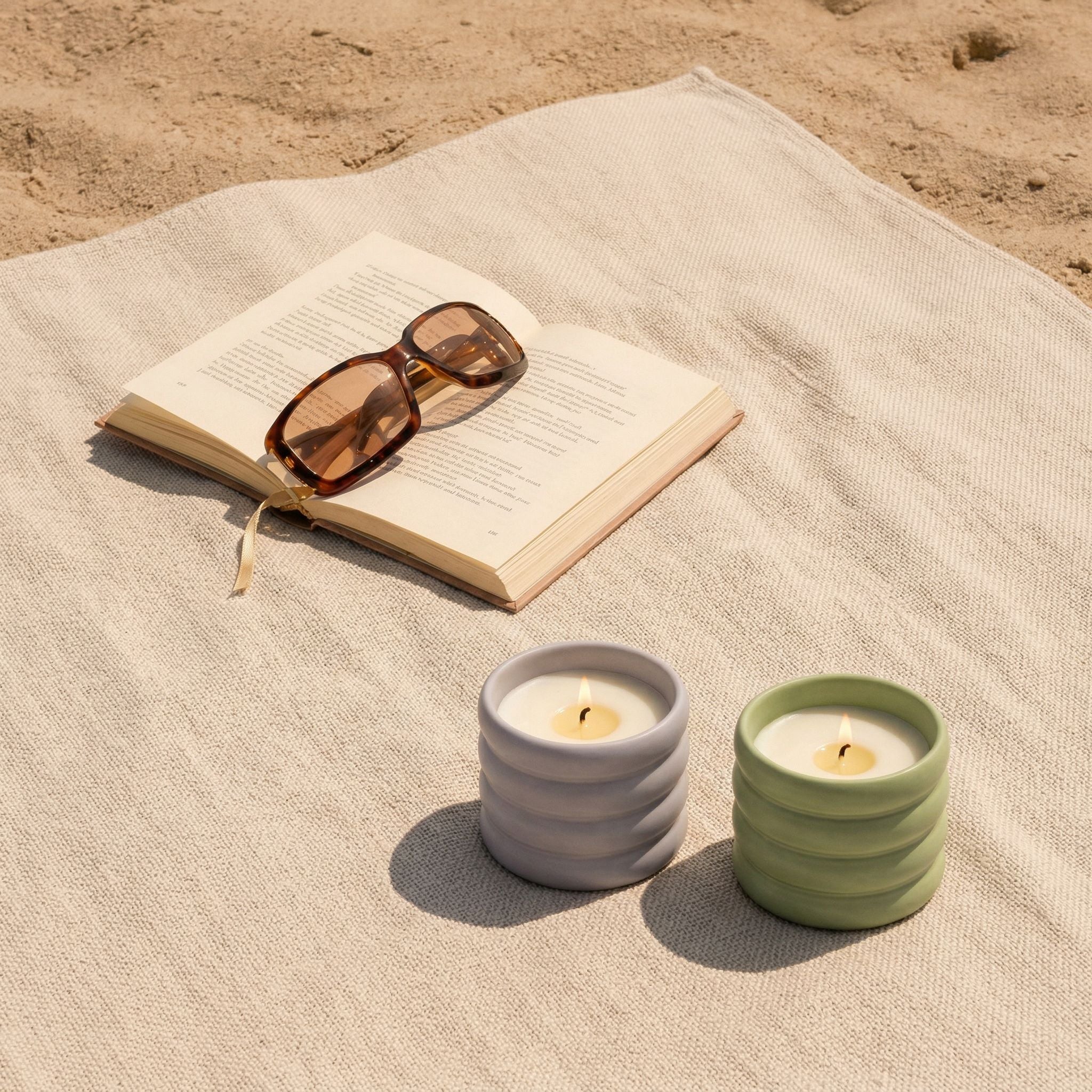Two candles on a sandy surface with an open book and sunglasses.