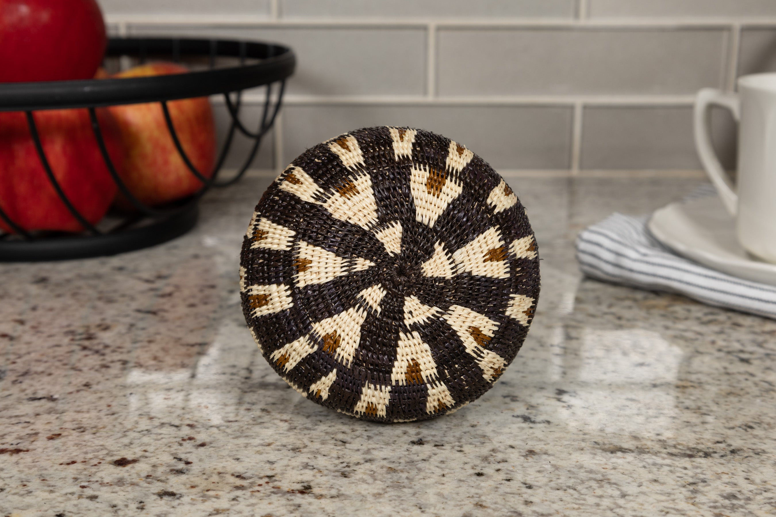 Decorative woven basket bottom on a kitchen counter with apples and a mug in the background.