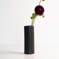 Black cylindrical vase with a single dark purple flower on a white background