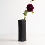 Black cylindrical vase with a single dark purple flower on a white background