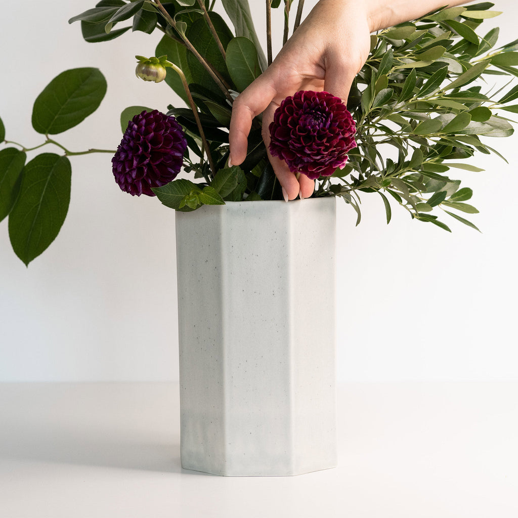 Hand arranging flowers in a white hexagonal vase on a light background