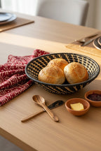 Bread rolls in a black woven basket on a wooden table with butter and jam.