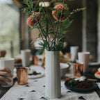 White vase with flowers on a table setting with plates and cups.