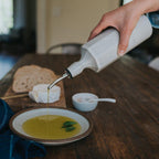Person pouring olive oil from a bottle onto a plate with bread and cheese on a wooden table.