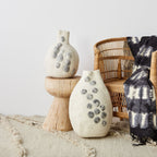 Two textured vases on a wooden stand with a wicker chair and tie-dye blanket in the background.