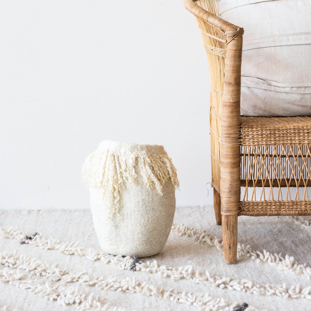 White textured vase with fringe on a light rug with a wicker chair in the background