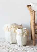 Two textured vases with fringe on a rug with a wicker chair in the background.