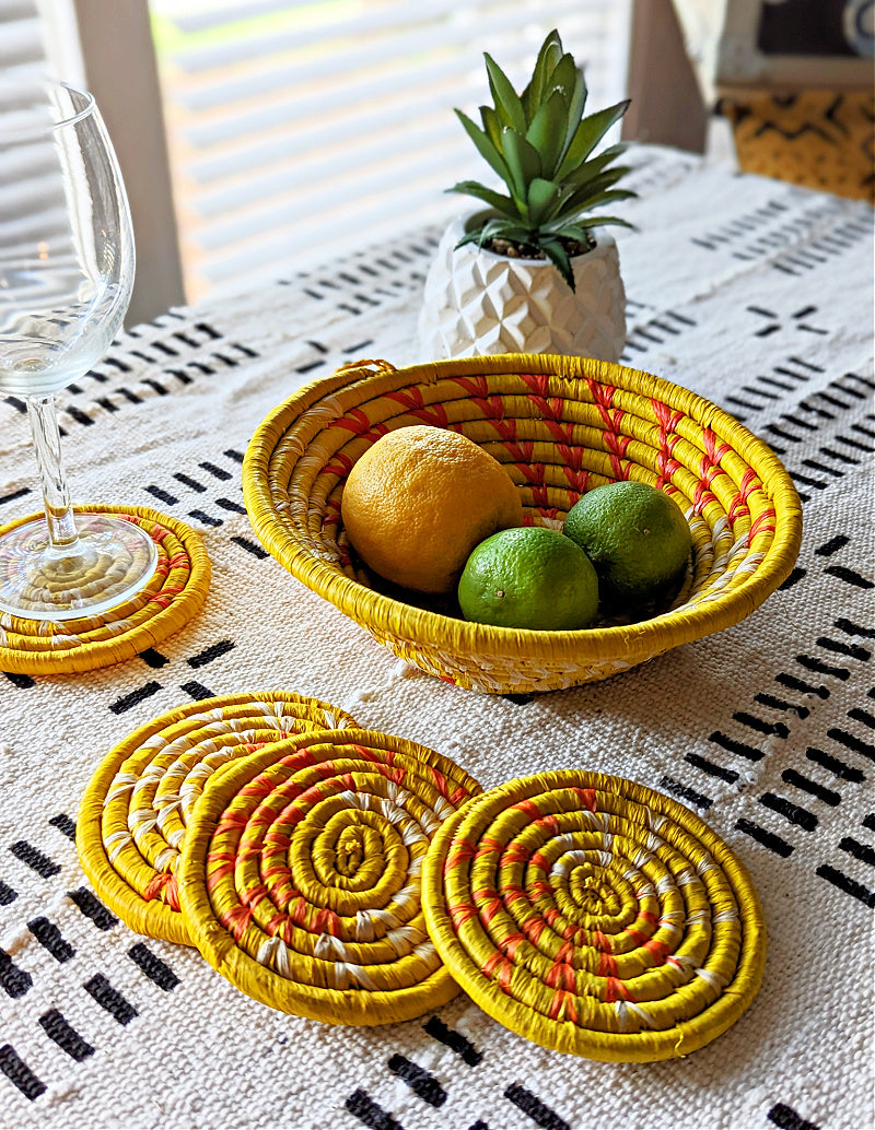 Yellow woven coasters on a table with a basket of fruit and a plant.