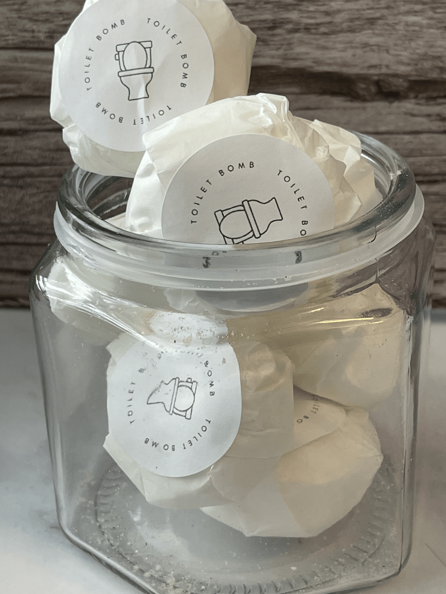 Glass jar filled with white toilet bombs labeled 'Toilet Bomb' on a wooden surface.