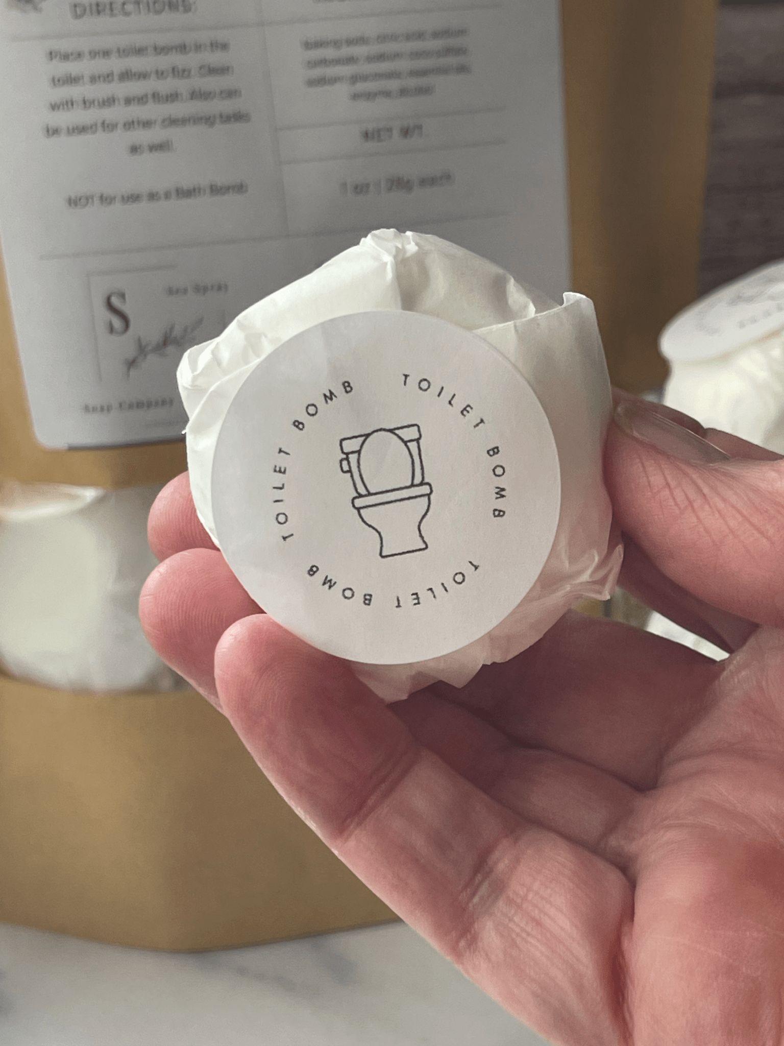 Hand holding a wrapped toilet bomb with a visible label against a blurred background