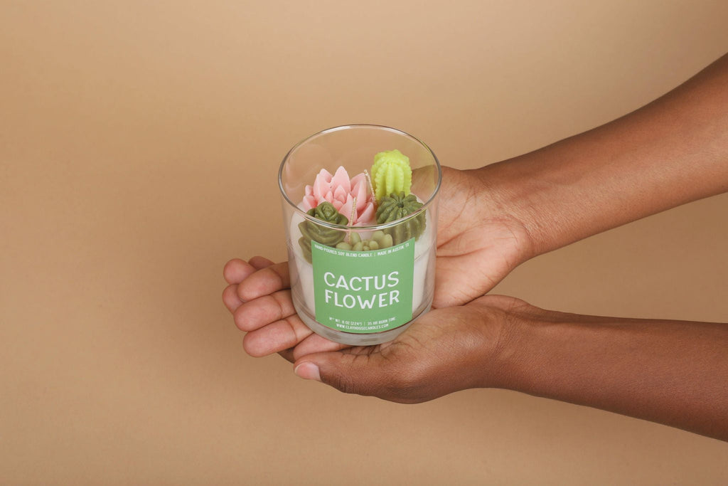 Hand holding a glass container with a cactus flower candle against a beige background
